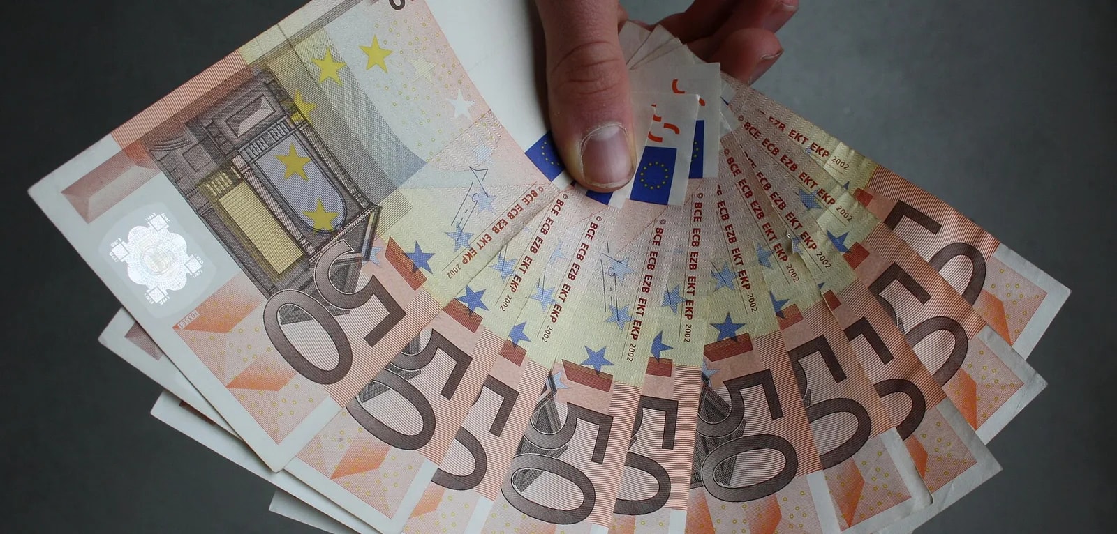 A hand fanning out a stack of crisp 50 euro banknotes against a dark background.
