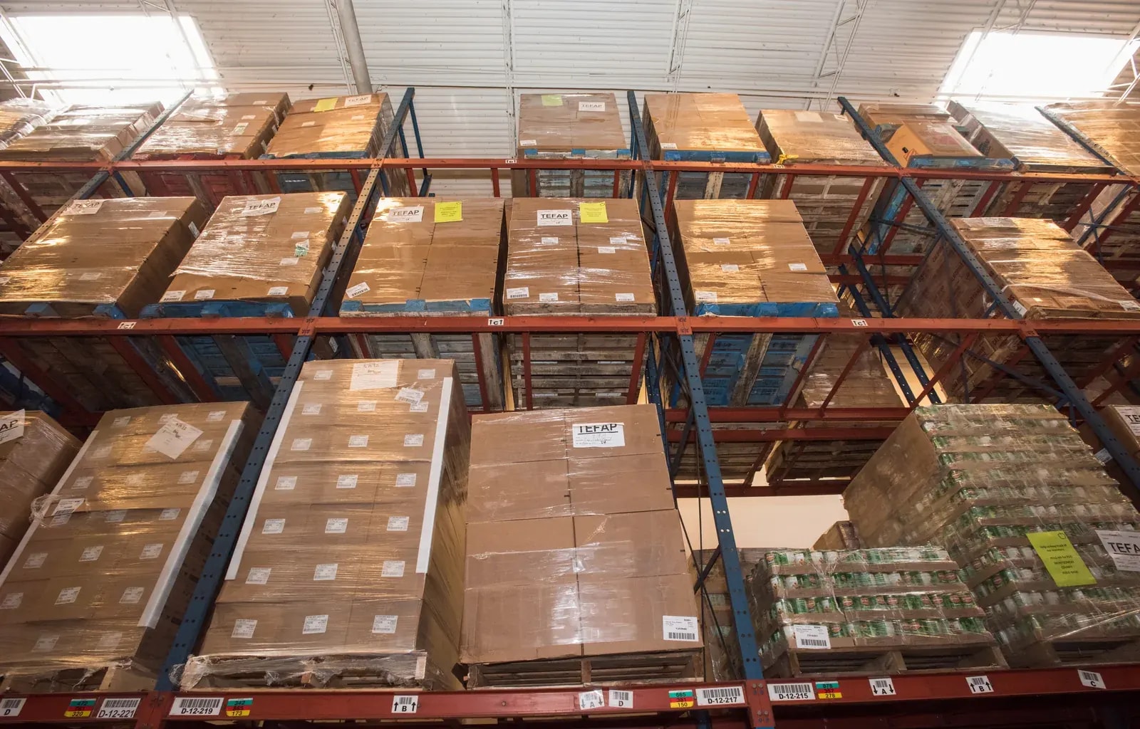 A warehouse aisle with tall red racking stacked with boxed and wrapped stock on pallets.